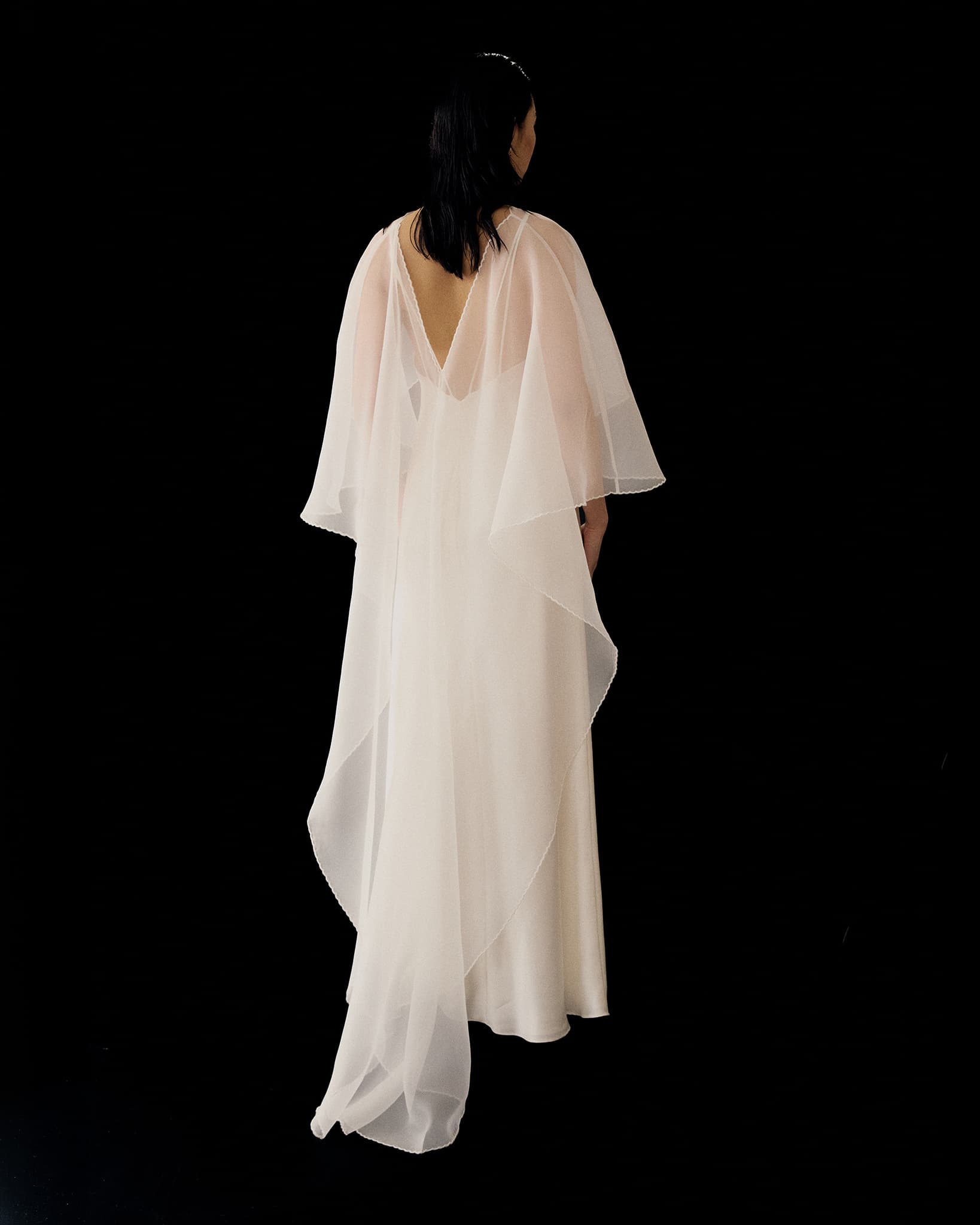 Against a black background, a person is shown from behind in the Noémiah Céline Dress—a long white evening gown with sheer, draped fabric flowing elegantly from the shoulders down the back.