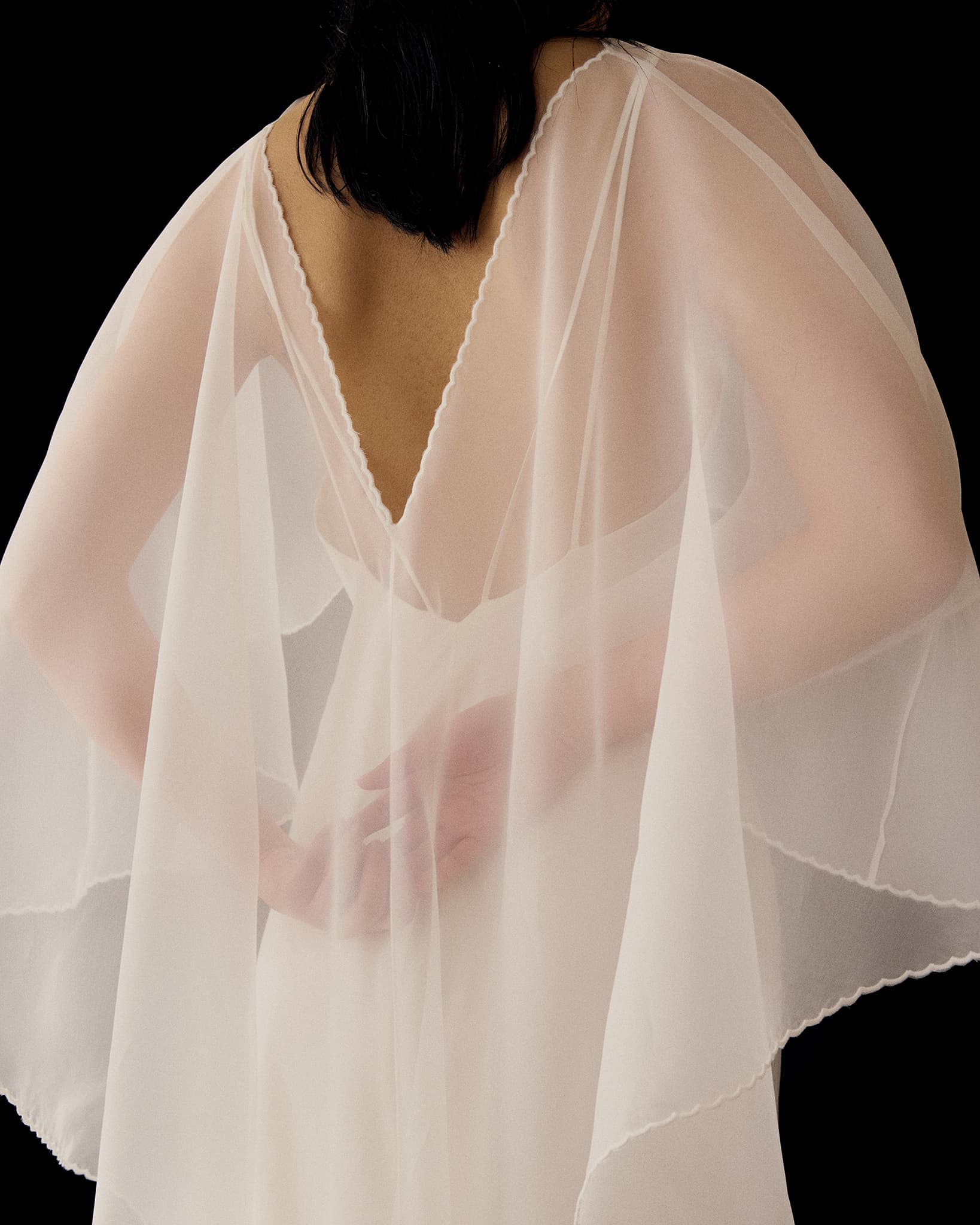 A person with dark hair stands with their back to the camera, wearing the sheer, flowing white Céline Dress by Noémiah with a deep V-shaped back. Their arms are crossed behind them against a solid black background.