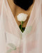 A person in the Noémiah Céline Dress, a sheer white bias cut style, holds a single white rose at their chest, its stem and leaves visible through the translucent fabric.