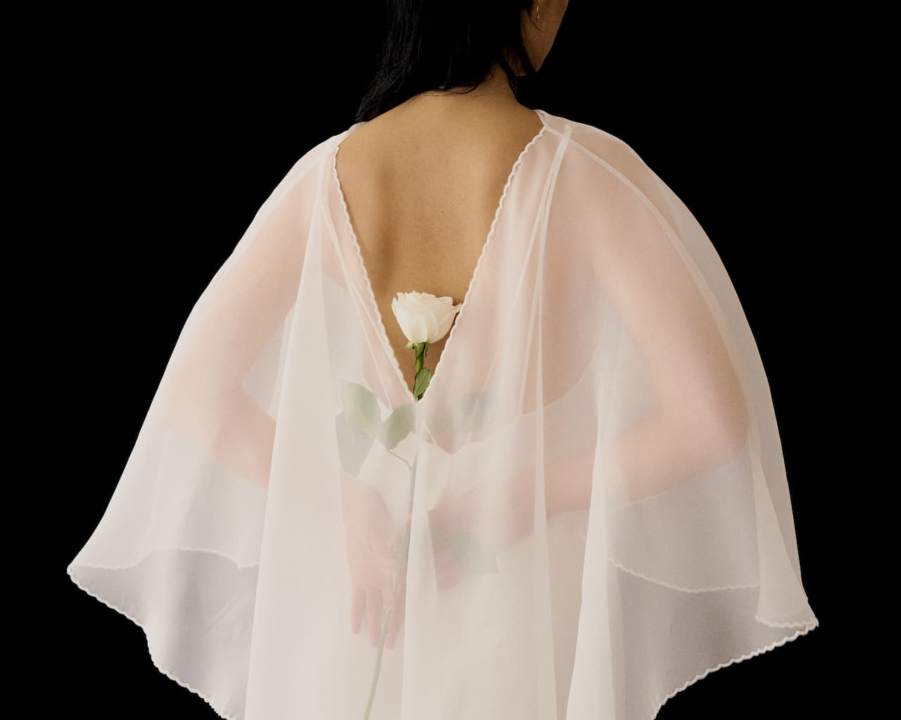 A person wearing the Céline Dress by Noémiah, made of sheer white fabric, stands with their back turned, holding a single white rose behind them against a black background.