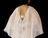 A person wearing the Céline Dress by Noémiah, made of sheer white fabric, stands with their back turned, holding a single white rose behind them against a black background.