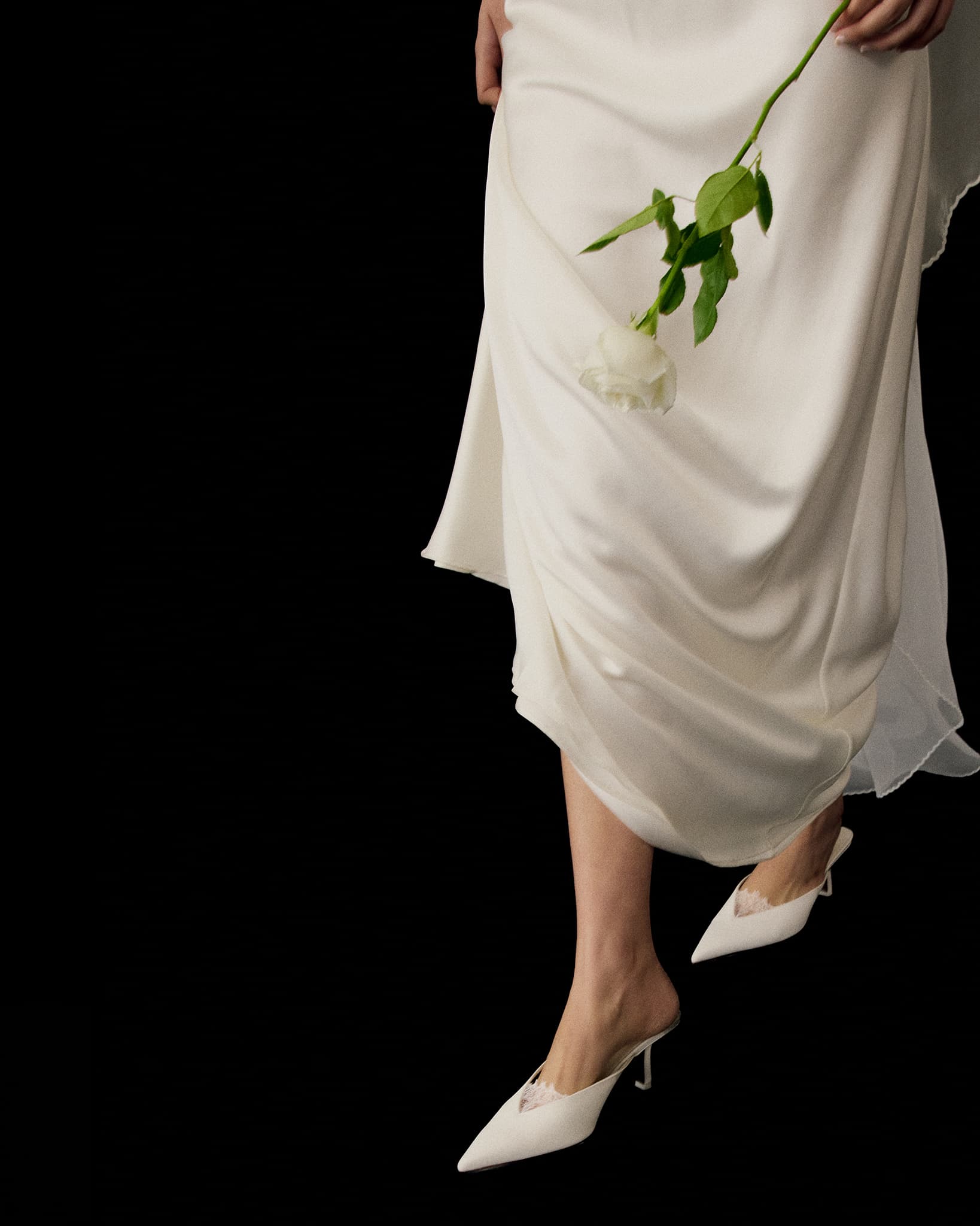 A person wears the Noémiah Céline Silk Satin Dress — Bridal Edit, holding a single white rose. Only their lower body is visible, paired with white pointed-toe heels, set against a striking black background.