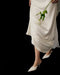 A person wears the Noémiah Céline Silk Satin Dress — Bridal Edit, holding a single white rose. Only their lower body is visible, paired with white pointed-toe heels, set against a striking black background.