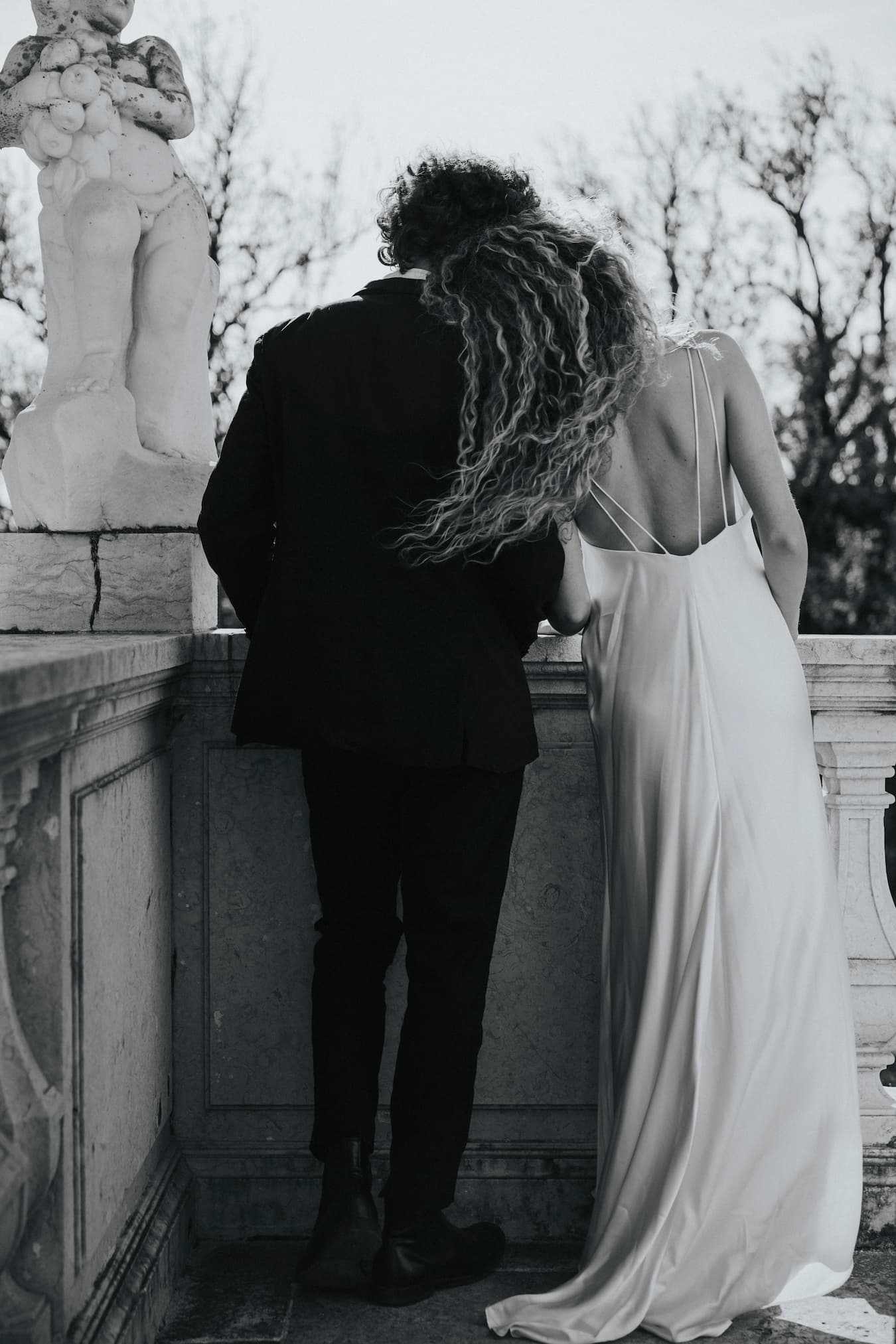 A couple stands closely on a balcony beside a stone railing and statue, the woman in a Noémiah Céline Silk Satin Dress — Bridal Edit leaning on the man's shoulder. They face away from the camera in this black-and-white image.