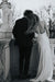 A couple stands closely on a balcony beside a stone railing and statue, the woman in a Noémiah Céline Silk Satin Dress — Bridal Edit leaning on the man's shoulder. They face away from the camera in this black-and-white image.