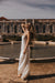 A woman in the Céline Silk Satin Dress — Bridal Edit by Noémiah stands barefoot by a fountain, basking in sunlight with her head tilted back and hand in her curls, ornate classical architecture behind her.