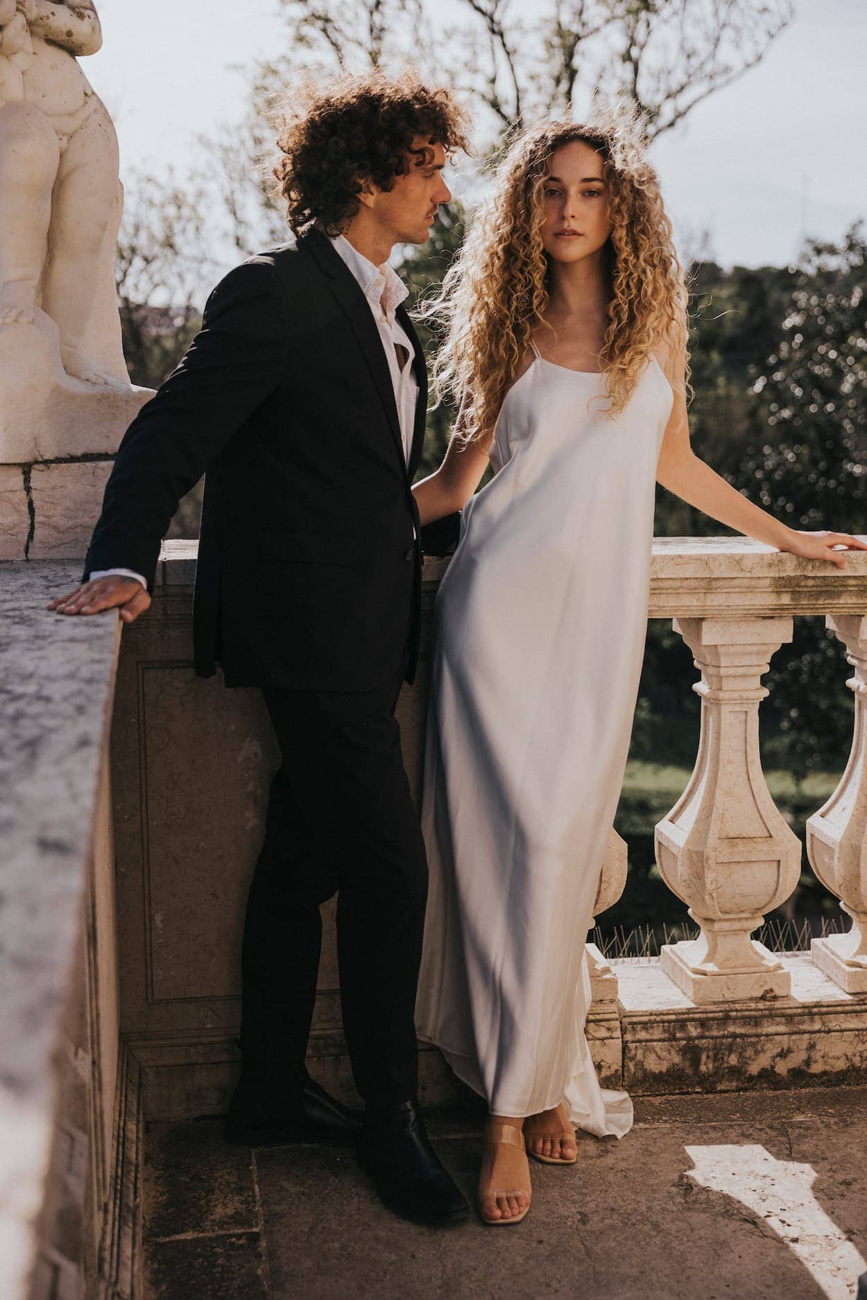 A man in a black suit and a woman in the Noémiah Céline Silk Satin Dress — Bridal Edit stand together on a stone balcony with ornate railings and greenery, both gazing ahead serenely.