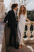 A man in a black suit and a woman in the Noémiah Céline Silk Satin Dress — Bridal Edit stand together on a stone balcony with ornate railings and greenery, both gazing ahead serenely.