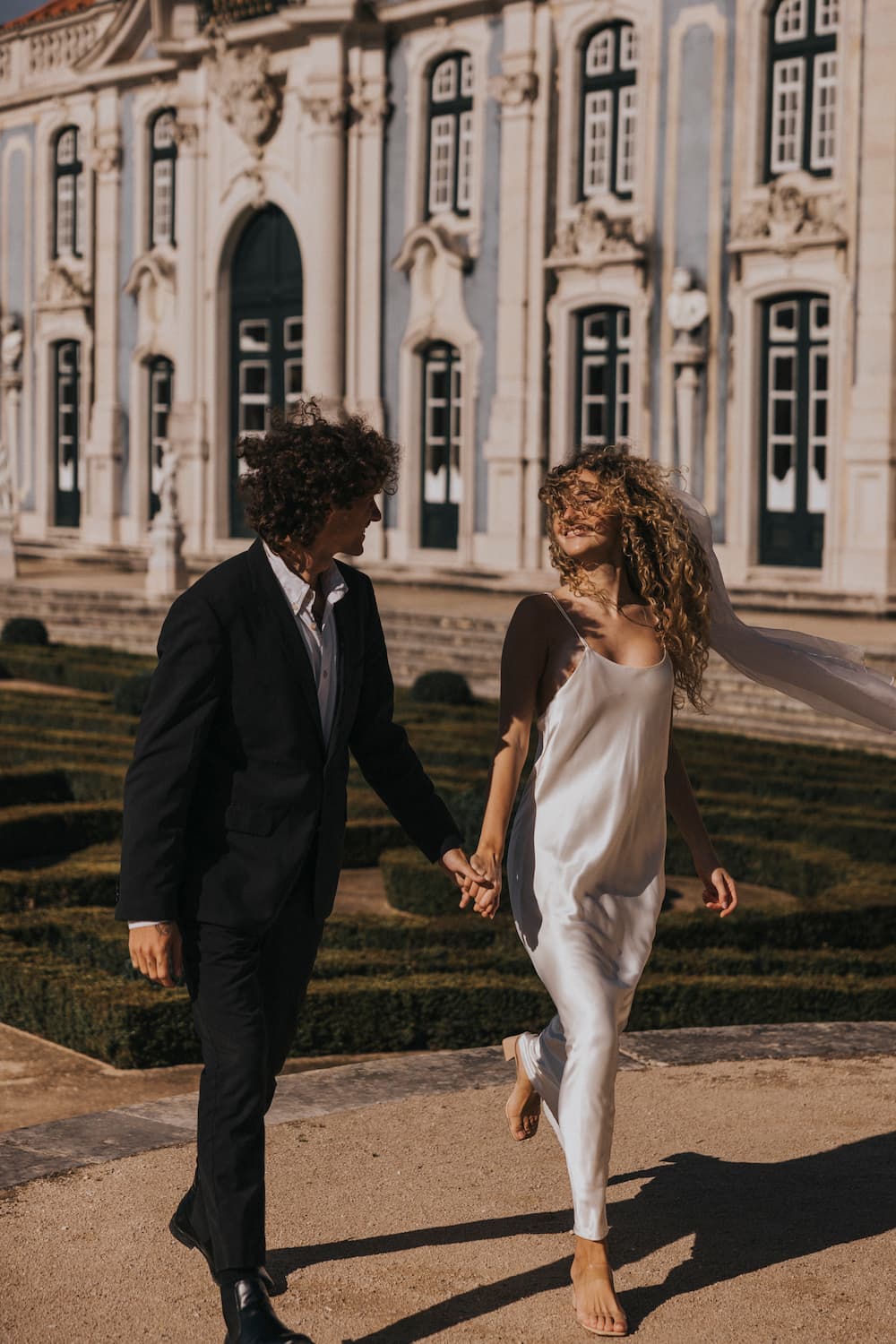 A barefoot bride wearing the Noémiah Céline Silk Satin Dress — Bridal Edit holds hands with a suited man as they smile and walk outside a grand building with neatly trimmed gardens.