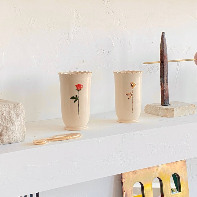 Two cream-colored, limited edition Noémiah Ceramic Vases with scalloped edges and painted flowers sit on a white shelf among minimalistic decor, including a stone block and an abstract metal sculpture.