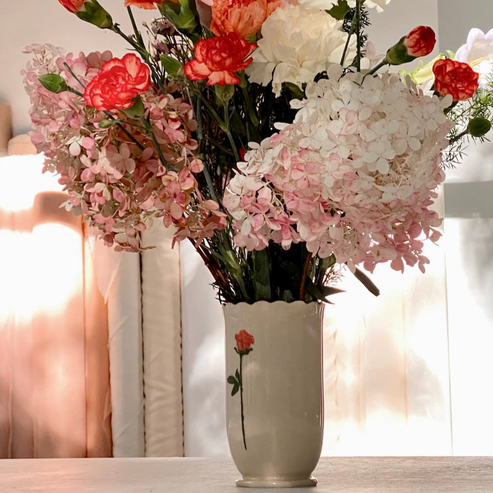The Noémiah Ceramic Vase, featuring a floral design and gold scalloped rim, holds pink carnations, white hydrangeas, and red flowers, displayed on a sunlit table.