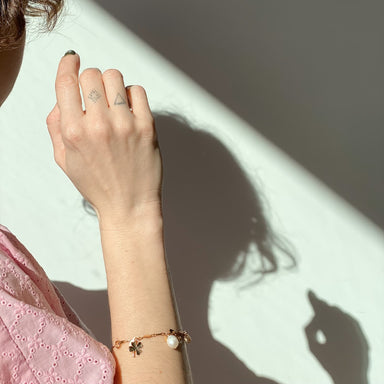 A woman's tattooed hand, adorned with the Noémiah Chance Bracelet—a gold-plated accessory featuring Swarovski crystals—casts a shadow against a sunlit wall. She wears a pink lace top, and the bracelet's charms include delicate freshwater pearls and a clover.