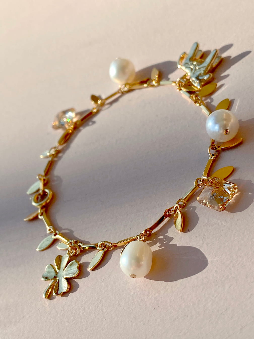 The Chance Bracelet by Noémiah, a gold-plated charm bracelet adorned with butterflies, leaves, freshwater pearls, and a cactus charm, is elegantly showcased on a light surface under the gentle glow of warm natural sunlight.