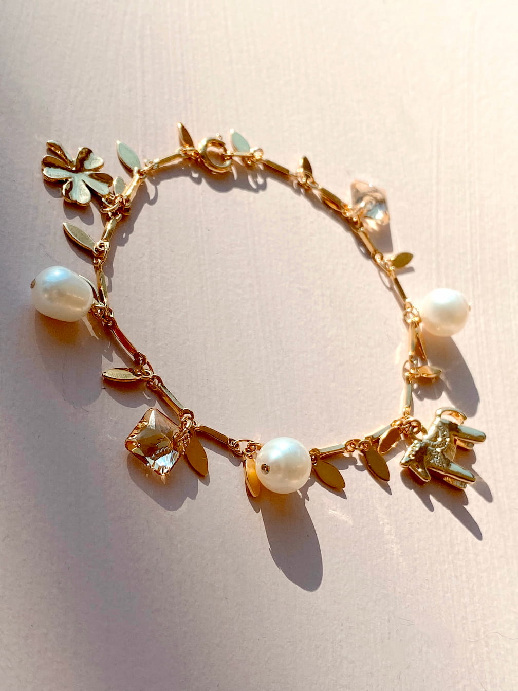 The Chance Bracelet by Noémiah is a delicate gold-plated piece featuring charms of leaves, a flower, freshwater pearls, and abstract shapes. It is beautifully displayed against a soft pink background with gentle shadows cast by the light.