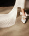 A person wearing white, pointed-toe shoes with straps stands on a smooth floor, while the Chapel Veil by Noémiah gracefully flows behind them, evoking elegance and movement against the neutral background.