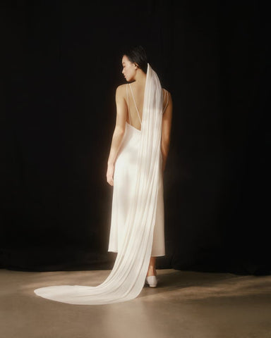 A woman in a white dress stands with her back to the camera against a dark background. Her Chapel Veil by Noémiah, crafted from hand-dyed ivory silk tulle, cascades gracefully to the floor, adding elegance to the scene. The lighting casts a soft glow on her and the metal tooth comb securing the veil.