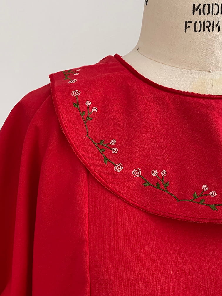 The Charlie Collared Dress by Noémiah is a red garment with a relaxed fit, featuring a large collar decorated with unique embroidery of delicate pink flowers and green leaves, beautifully showcased on a dress form.