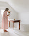 A woman wearing the relaxed fit, light pink Charlie Collared Dress by Noémiah stands in a white room, holding a vase filled with colorful flowers next to a wooden table. The table holds a small fruit bowl. Soft natural light streams through a window, creating a peaceful atmosphere that highlights the dress's unique embroidery.