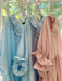 Three Charlie Collared Dresses by Noémiah, in pastel shades—two light blue and one pink—are elegantly displayed on hangers. Each dress boasts distinct embroidery on the collar and ruffled sleeves. Their relaxed fit is showcased against a backdrop of green foliage visible through a window.