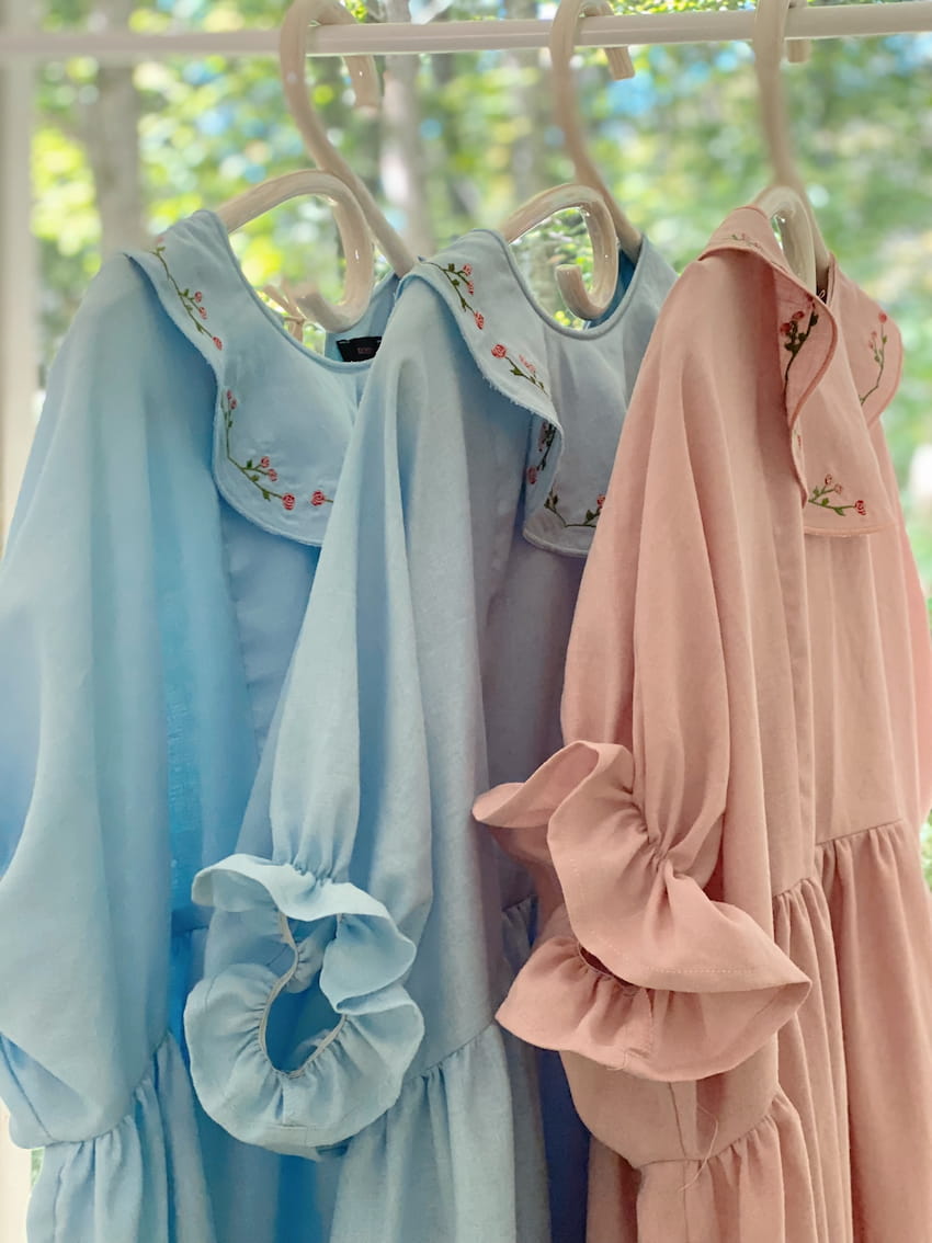 Three Charlie Collared Dresses by Noémiah, in pastel shades—two light blue and one pink—are elegantly displayed on hangers. Each dress boasts distinct embroidery on the collar and ruffled sleeves. Their relaxed fit is showcased against a backdrop of green foliage visible through a window.