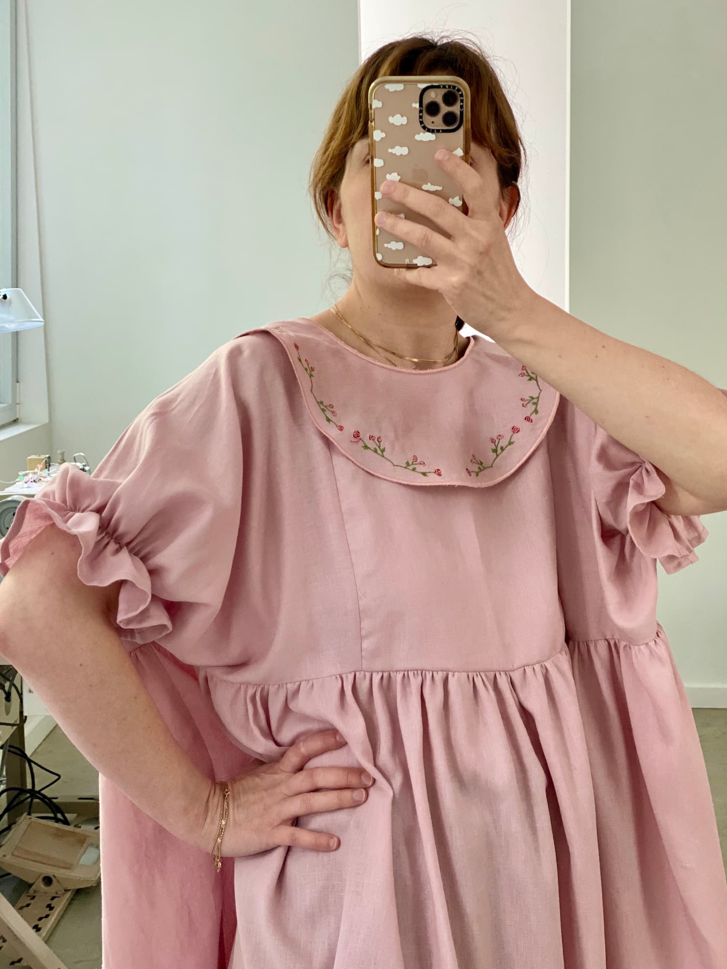 In a bright and airy room featuring a window and some furniture, a person takes a mirror selfie while wearing the relaxed-fit Charlie Collared Dress by Noémiah. The dress is pink and adorned with unique floral embroidery on the collar. Their phone case displays a cloud pattern.