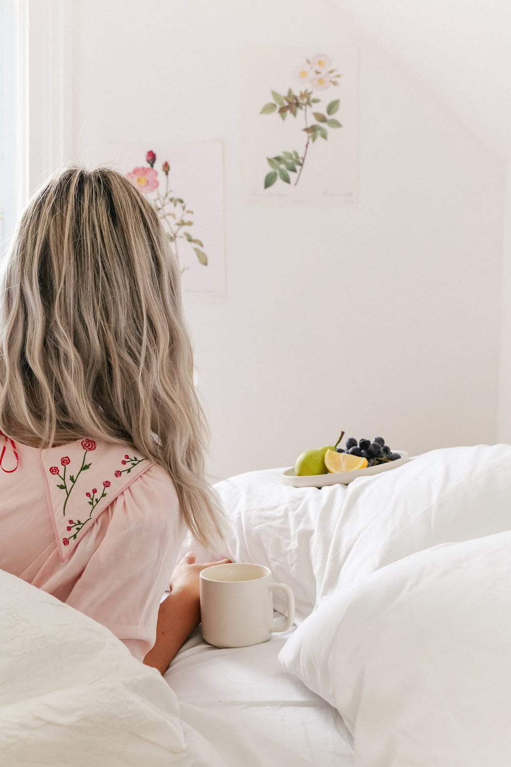 A woman with long, blonde hair sits on a bed with white sheets, wearing a relaxed fit Charlie Collared Dress by Noémiah. She holds a mug and faces floral artwork on a white wall. A bedside table showcases distinctive embroidery next to a plate of grapes, lemons, and a pear.