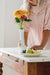 A person in a relaxed-fit Charlie Collared Dress by Noémiah arranges a white vase with yellow and pink flowers on a table. Beside it, a plate with sliced fruit and grapes adds to the inviting scene in the bright room filled with natural light.