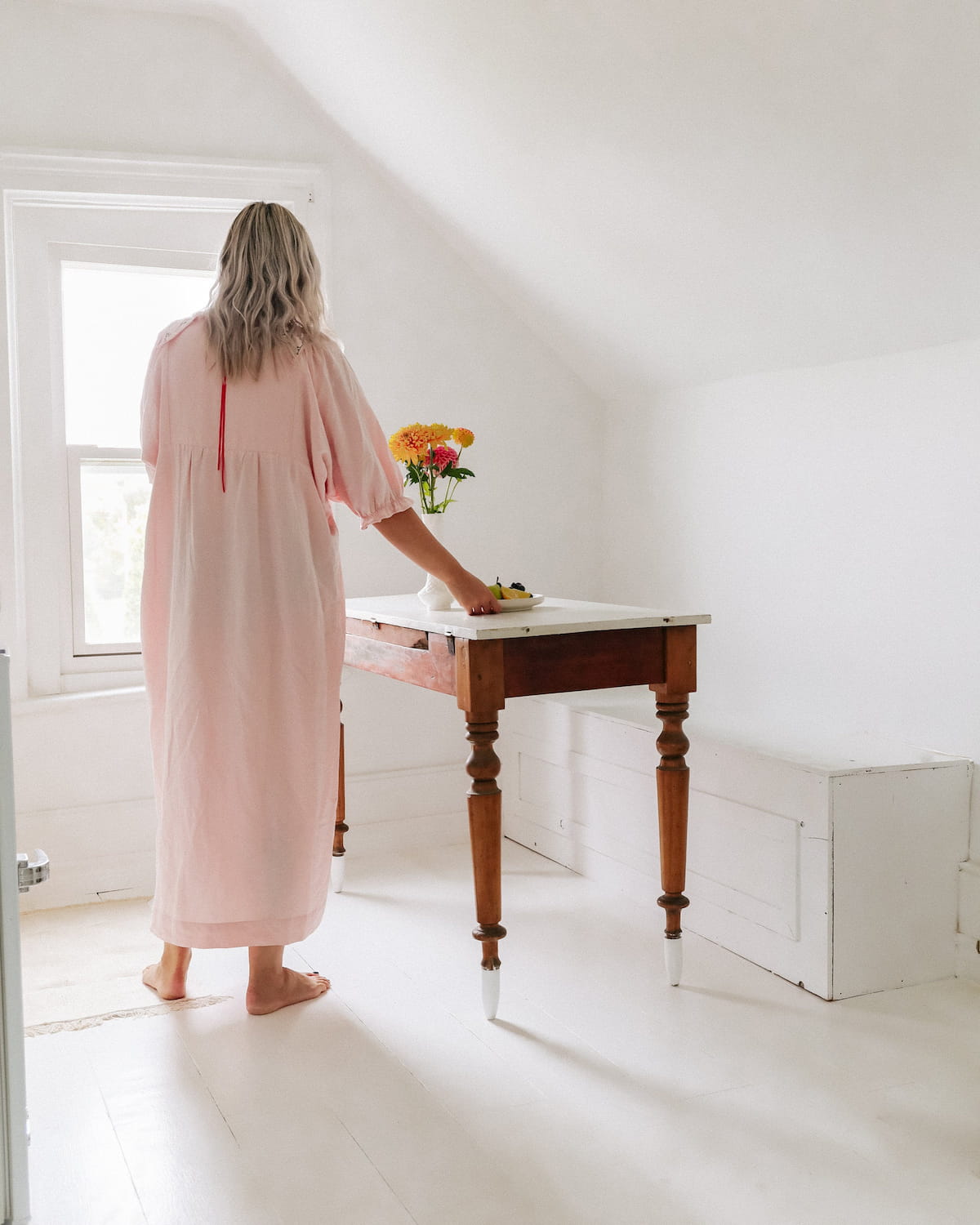 A woman in a long pink Charlie Collared Dress by Noémiah stands barefoot in a bright room, arranging flowers on a wooden table by the window. The space is minimalistic with white floors and walls, accentuating the dress's unique embroidery that adds charm to her serene setting.