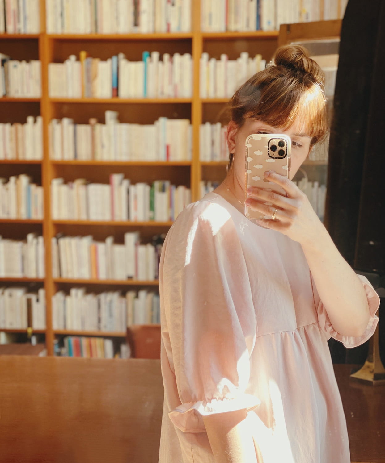 A person wearing the Charlie Collared Dress by Noémiah, featuring unique embroidery, takes a mirror selfie with their phone, covering most of their face. Their hair is tied up in a bun, and in the background, a large bookshelf filled with books can be seen.