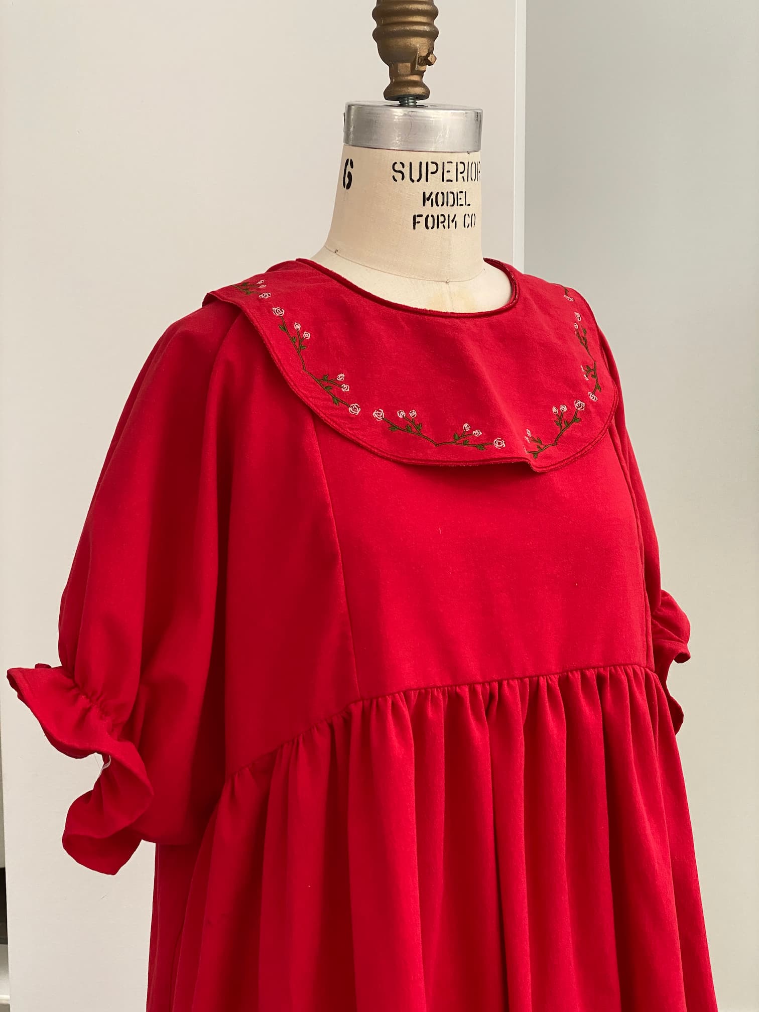 The Charlie Collared Dress by Noémiah boasts a relaxed fit in a striking red design, highlighted by a gathered waist and puffed sleeves on display. Its wide collar is adorned with distinctive floral embroidery, and the sleeves are elegantly tied with matching fabric bows.