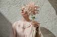 A person with short blond hair and closed eyes stands against a textured wall, holding a large, pale pink flower in front of their face. They are wearing the light Noémiah Charlie Collared Dress, featuring unique floral embroidery on the collar. Sunlight casts soft shadows.