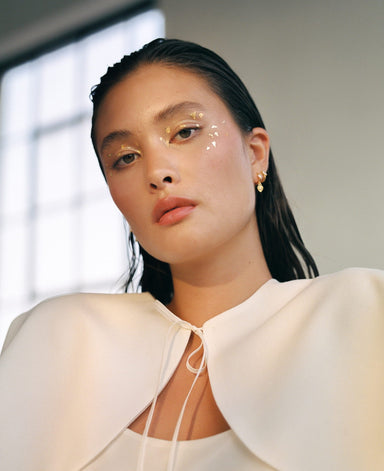 The individual, with wet hair slicked back, wears a white outfit and the Charlie Earrings by Noémiah as they gaze at the camera. Gold flecks add decoration near their eyes, and a window bathes the scene in soft light from behind.