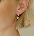 A close-up of a person's ear showcases the Charlie Earrings by Noémiah, lightweight hoops featuring a charming double heart design. Above the hearts, a small clear stone provides a touch of sparkle. The person's short, light-colored hair beautifully accentuates this elegant accessory.