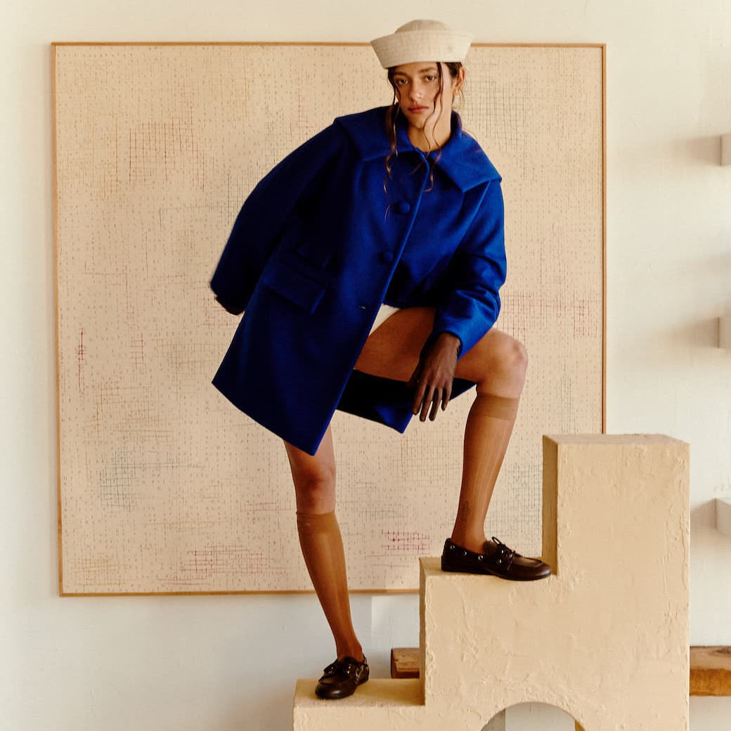 A person wearing the Noémiah Charlot Coat — Classic, a white beret, and dark shoes poses with one leg on a beige, staircase-shaped structure in a minimal, modern room with art and shelves in the background.