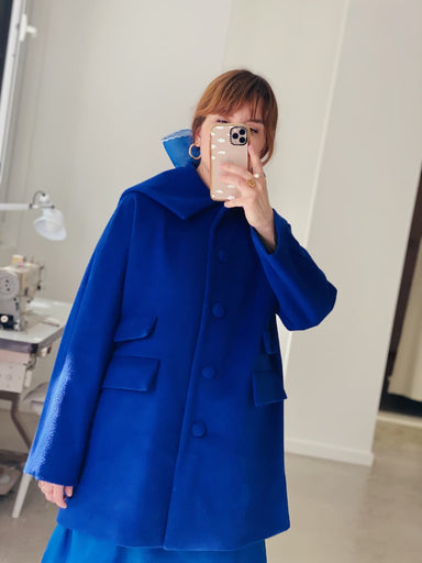 A person takes an indoor mirror selfie wearing the Noémiah Charlot Coat — Classic in bright blue. Large buttons and flap pockets are visible, with their face partly obscured by a smartphone and a sewing machine in the background.