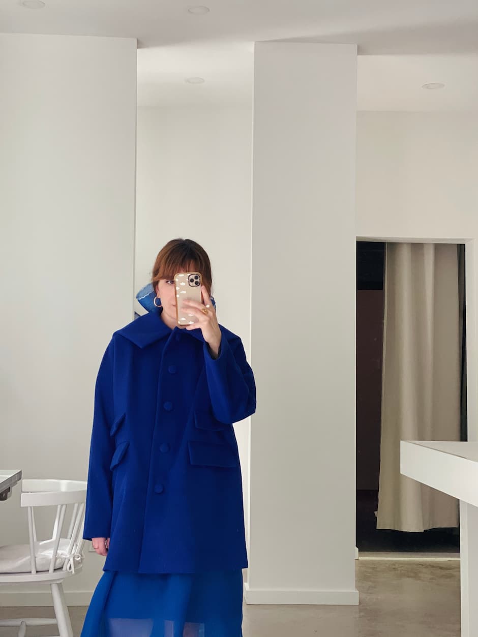 A person stands indoors taking a mirror selfie, wearing the Noémiah Charlot Coat — Classic in bright blue with a matching skirt. White walls, a chair, and a curtained doorway enhance the outfit’s modern silhouette.