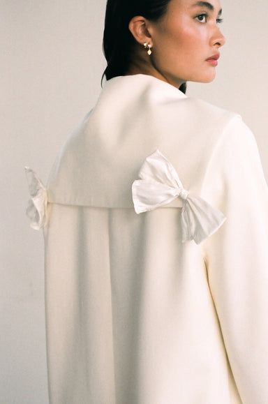 A woman stands against a plain background wearing the Charlot Coat by Noémiah. This cream sailor coat features silk taffeta bows on the back, highlighting its unique design as she looks to the side. Her dark hair is tied back, and she accessorizes with small hoop earrings.