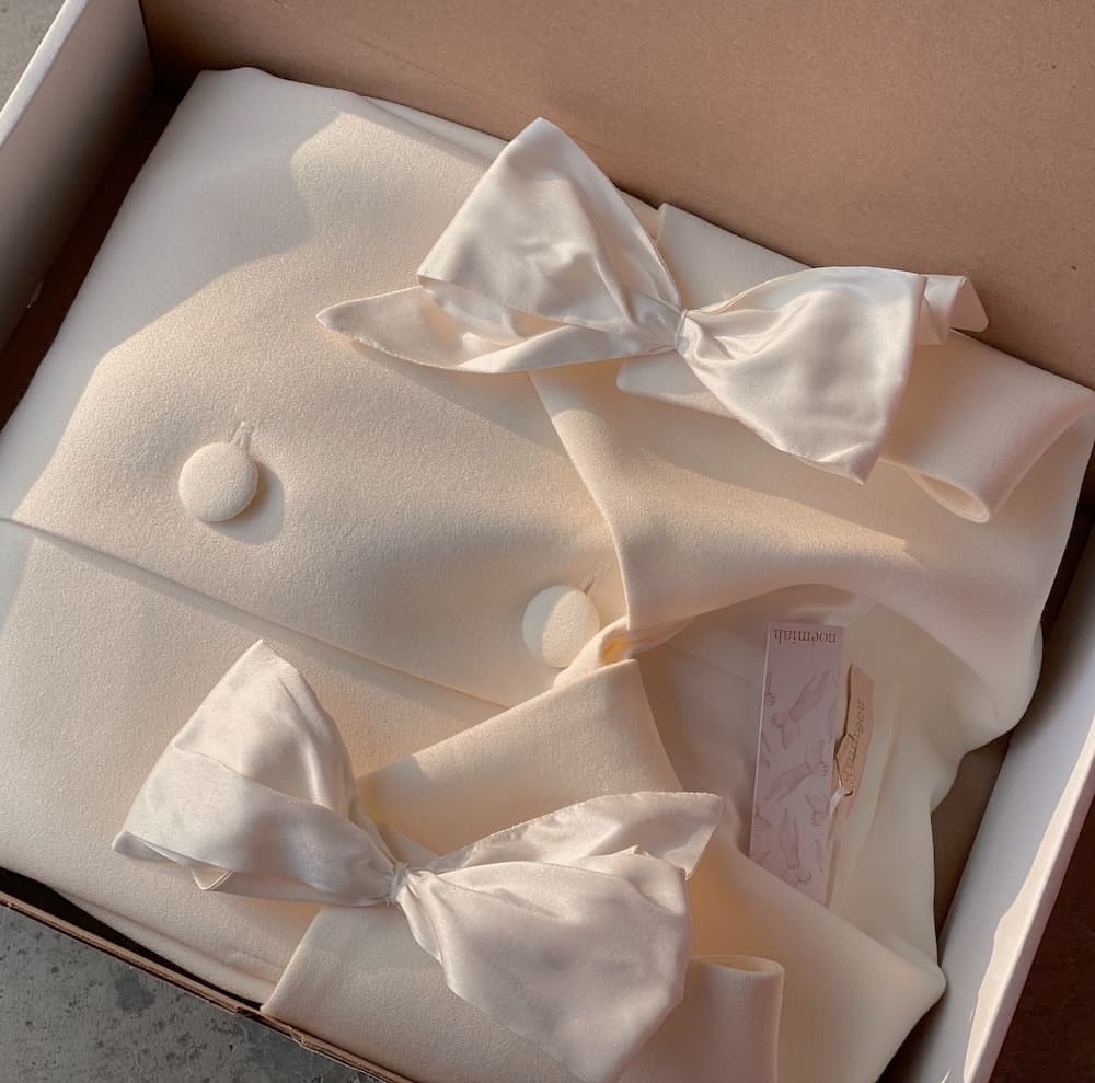 A neatly folded Charlot Coat from the brand Noémiah, featuring elegant buttons and two silk taffeta bows, is carefully placed in a cardboard box. A pink tag with text is tucked between the folds, lit gently by soft light.