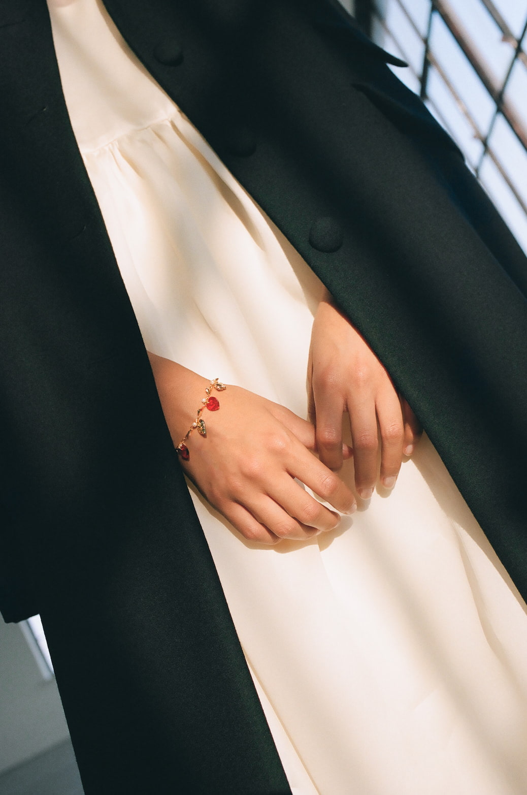 A close-up captures a person elegantly dressed in a white outfit paired with the Noémiah Charlot Coat, their hands adorned with a gold bracelet featuring small red charms. The softly lit background enhances the elegance of the attire and accessory.