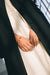 A close-up captures a person elegantly dressed in a white outfit paired with the Noémiah Charlot Coat, their hands adorned with a gold bracelet featuring small red charms. The softly lit background enhances the elegance of the attire and accessory.