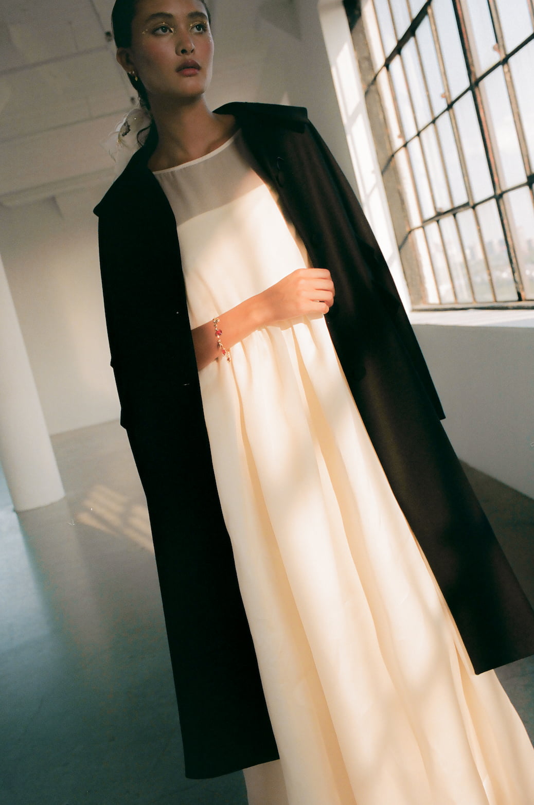 A woman stands indoors wearing a flowing cream dress adorned with silk taffeta bows, draping the Charlot Coat by Noémiah over her shoulders. Positioned near large industrial-style windows, natural light fills the space as she gazes toward the camera.