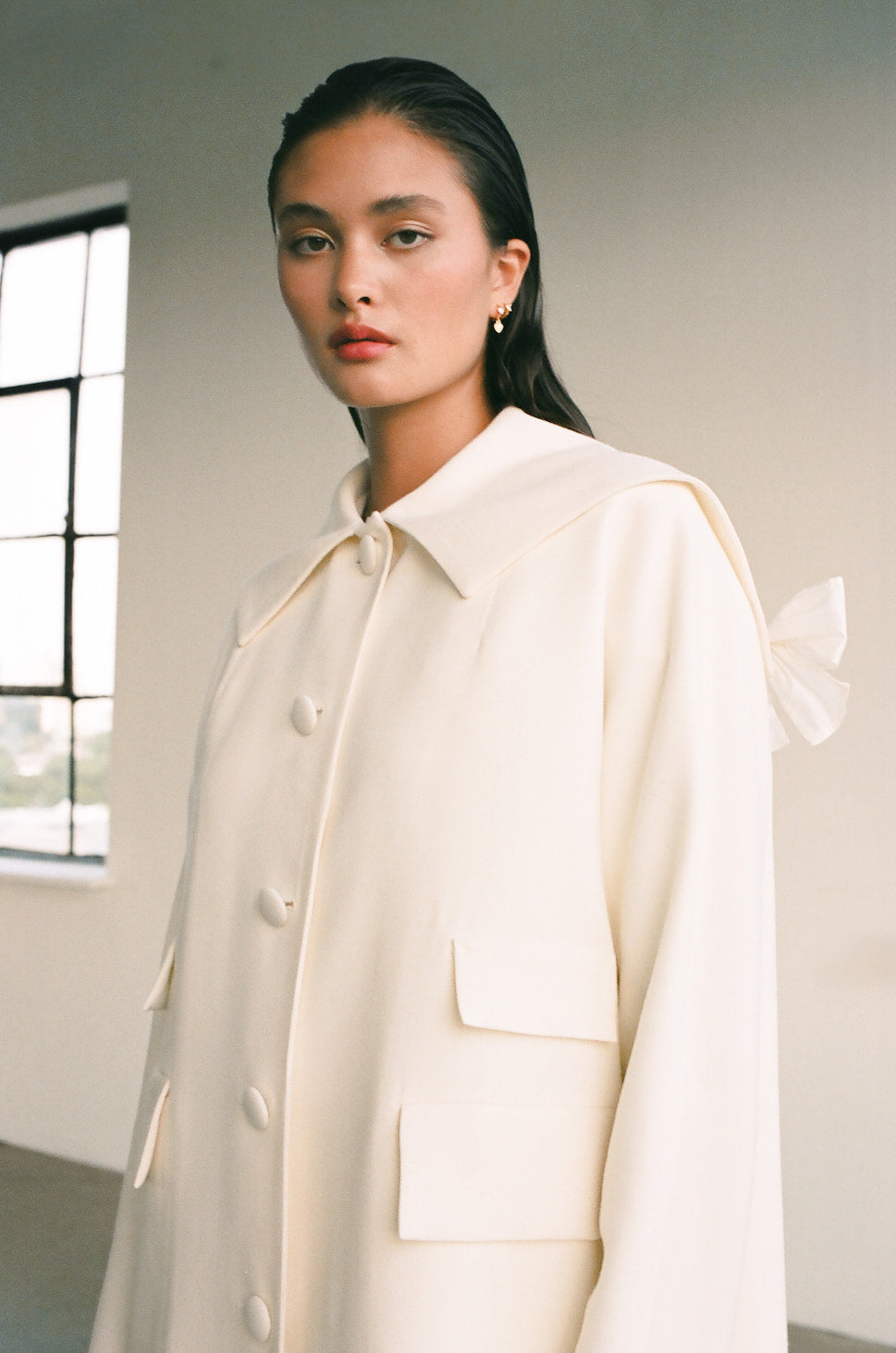 A person wearing the Charlot Coat by Noémiah poses indoors near a window, looking toward the camera. This cream-colored sailor coat made from wool crepe showcases a buttoned front and large pockets. The neutral-tone setting enhances the elegant look of the outfit.