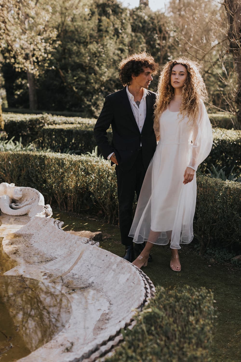 A man in a black suit and a woman wearing the Charlotte Dress from Noémiah's Bridal Edit stand by a quiet fountain in the garden. The dress, crafted from 100% silk organza, flows elegantly as they gaze into the distance, surrounded by lush greenery and sunlight.