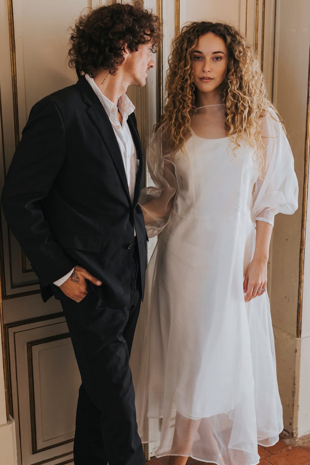 A man in a black suit stands beside a woman wearing the Bridal Edit Charlotte Dress by Noémiah, made from 100% silk organza. Both individuals have curly hair and are positioned in front of an ornate, light-colored wall, exuding a formal yet non-traditional bridal elegance.