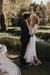 In a lush garden, a couple stands closely together, exuding a romantic ambiance. The woman is elegantly dressed in the flowing Charlotte Dress — Bridal Edit by Noémiah, crafted from 100% silk organza. The man complements her look in a dark suit as they gaze affectionately at each other, perfectly capturing the essence of non-traditional brides amidst the greenery with sunlight filtering through the trees.