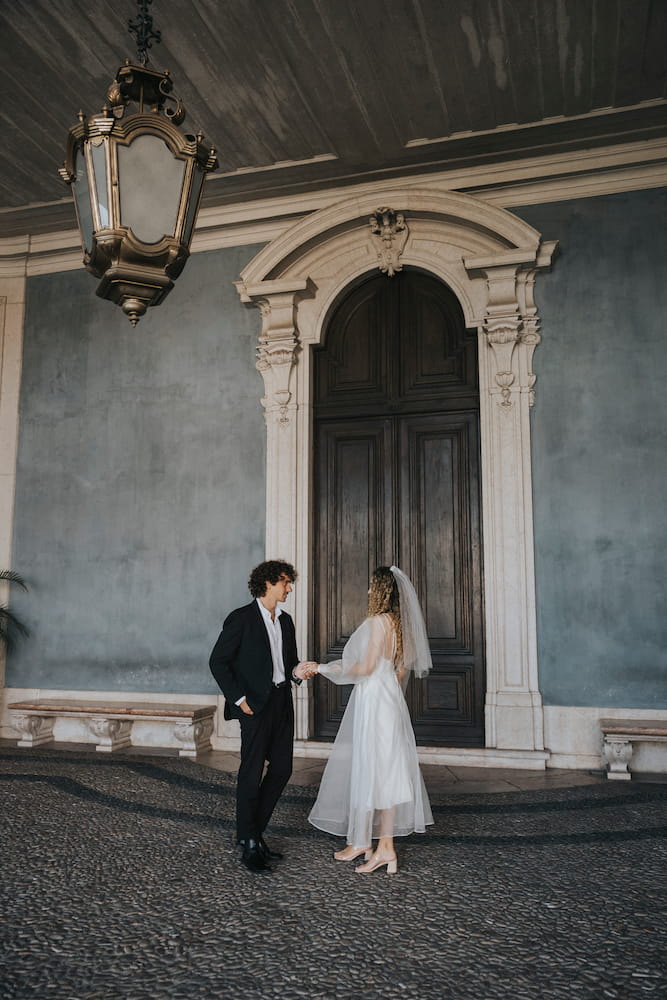 A couple stands in front of a large, ornate wooden door. The man is in a black suit, while the woman captivates in a Charlotte Dress — Bridal Edit by Noémiah, made from 100% silk organza. They are holding hands on a cobblestone floor under an elegant hanging lantern, embodying the grace of non-traditional brides.