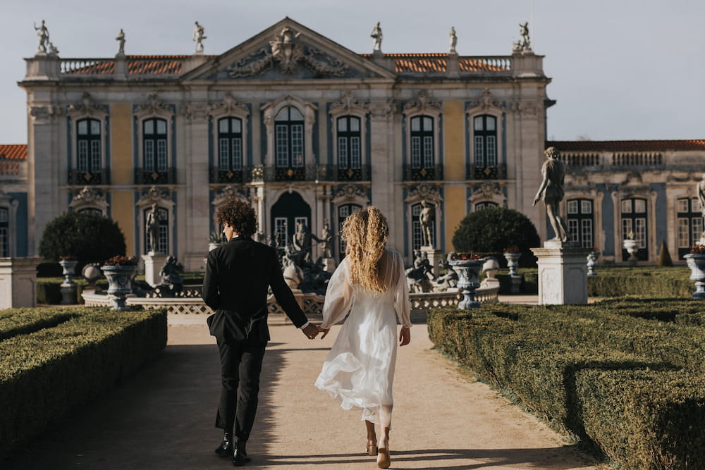 A couple walks hand in hand towards an ornate, historic building surrounded by a manicured garden. The woman, a non-traditional bride, is elegantly dressed in the flowing Charlotte Dress from the Bridal Edit by Noémiah, crafted entirely from 100% silk organza, while the man wears a black suit. Statues and trimmed hedges line their path.
