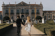 A couple walks hand in hand towards an ornate, historic building surrounded by a manicured garden. The woman, a non-traditional bride, is elegantly dressed in the flowing Charlotte Dress from the Bridal Edit by Noémiah, crafted entirely from 100% silk organza, while the man wears a black suit. Statues and trimmed hedges line their path.
