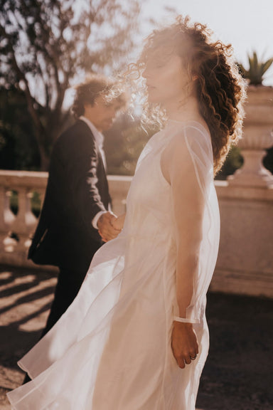 A couple holds hands under the sun. The woman, a non-traditional bride wearing Noémiah's Charlotte Dress — Bridal Edit, crafted from 100% silk organza, faces the camera as sunlight illuminates her curly hair. The man in a suit follows behind, set against a softly focused garden or park background.