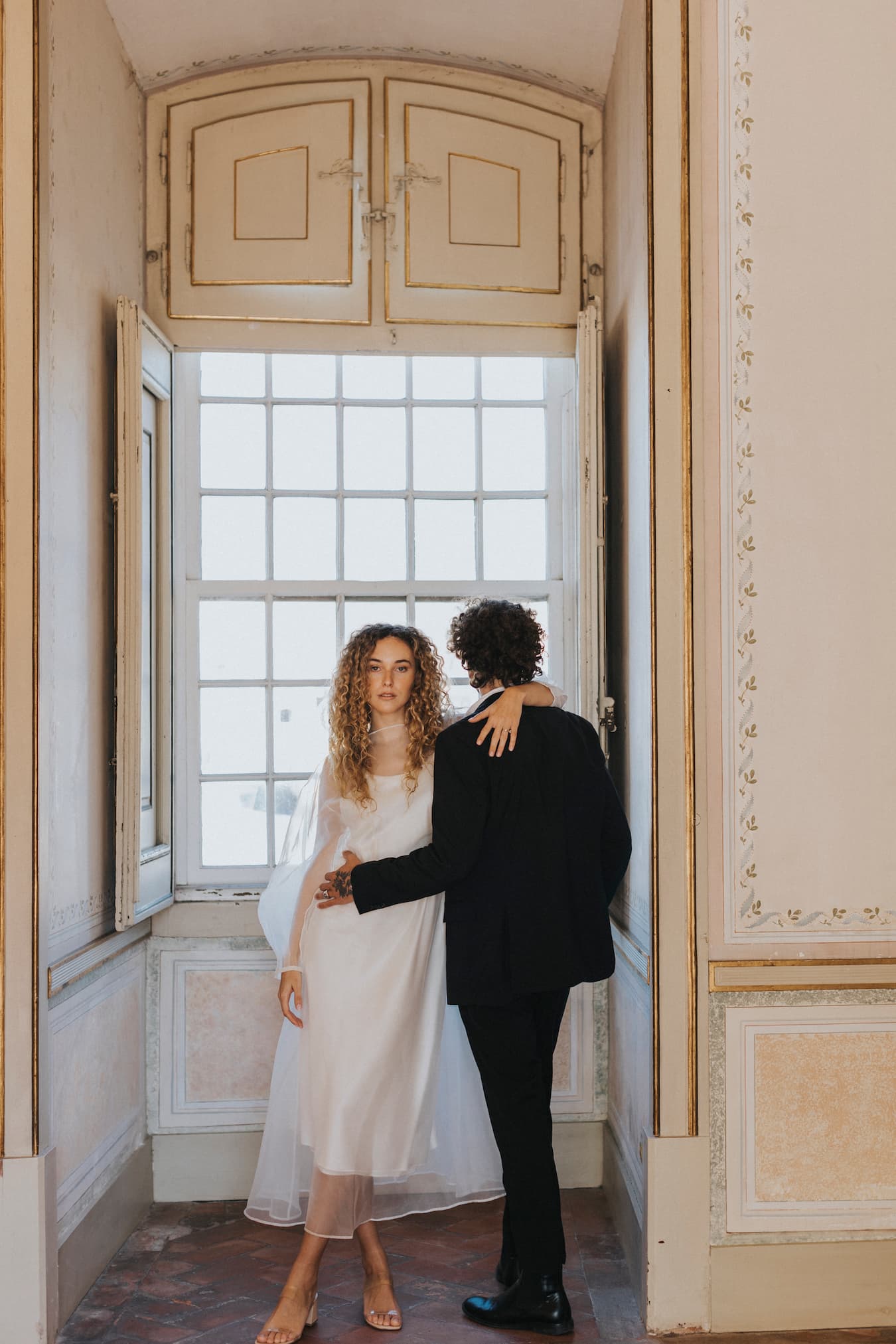 In an elegant room with ornate trim and soft lighting, a couple stands intimately in front of a large window. The woman, wearing Noémiah's Charlotte Dress from their Bridal Edit collection, crafted from 100% silk organza, gazes directly at the camera. Meanwhile, the man dressed in a dark suit faces away.